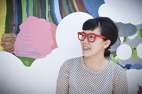 Female student with red-rmmed glasses sits in front of colorful artwork.